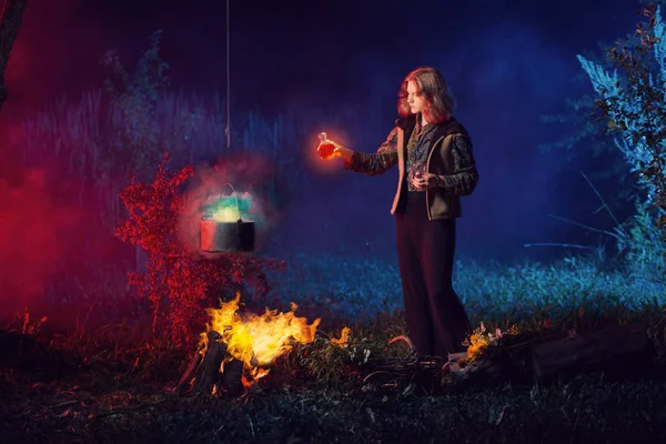 young witch by  fire in night forest prepares  magic potion