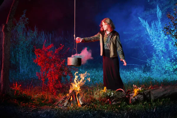 Joven bruja por el fuego en el bosque nocturno prepara poción mágica — Foto de Stock
