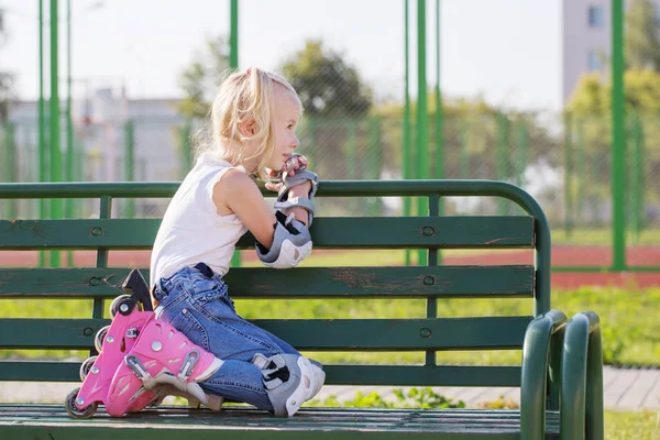 Маленькая девочка сидит на скамейке на детской площадке в роликовых коньках — стоковое фото