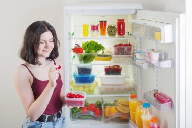 Kirazlı gülümseyen kız sağlıklı yiyecekli açık buzdolabının yanında.