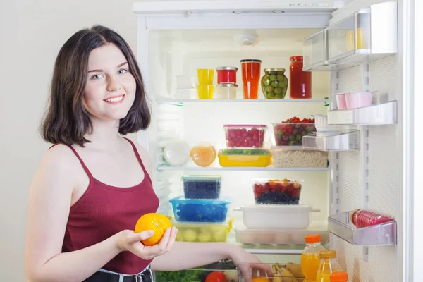 在开着的冰箱旁边放着橙子的微笑女孩 — 图库照片