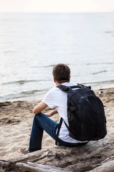 バックパックを持つ若い男が海辺でログの上に座る — ストック写真