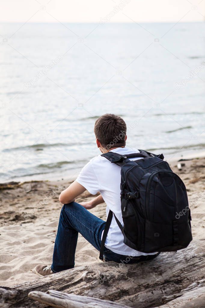 Young Man with Backpack sit on the Log at Seaside