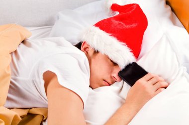 Yorgun genç adam Noel Baba şapkası içinde uyku bir telefonla yatakta