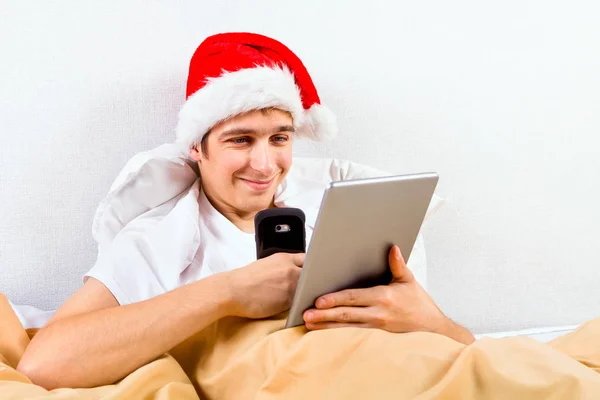 Jonge Man Kerstmuts Met Een Tabletcomputer Een Telefoon Het Bed — Stockfoto