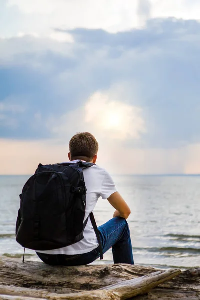 バックパックを持つ孤独な若い男が海辺でログの上に座る — ストック写真