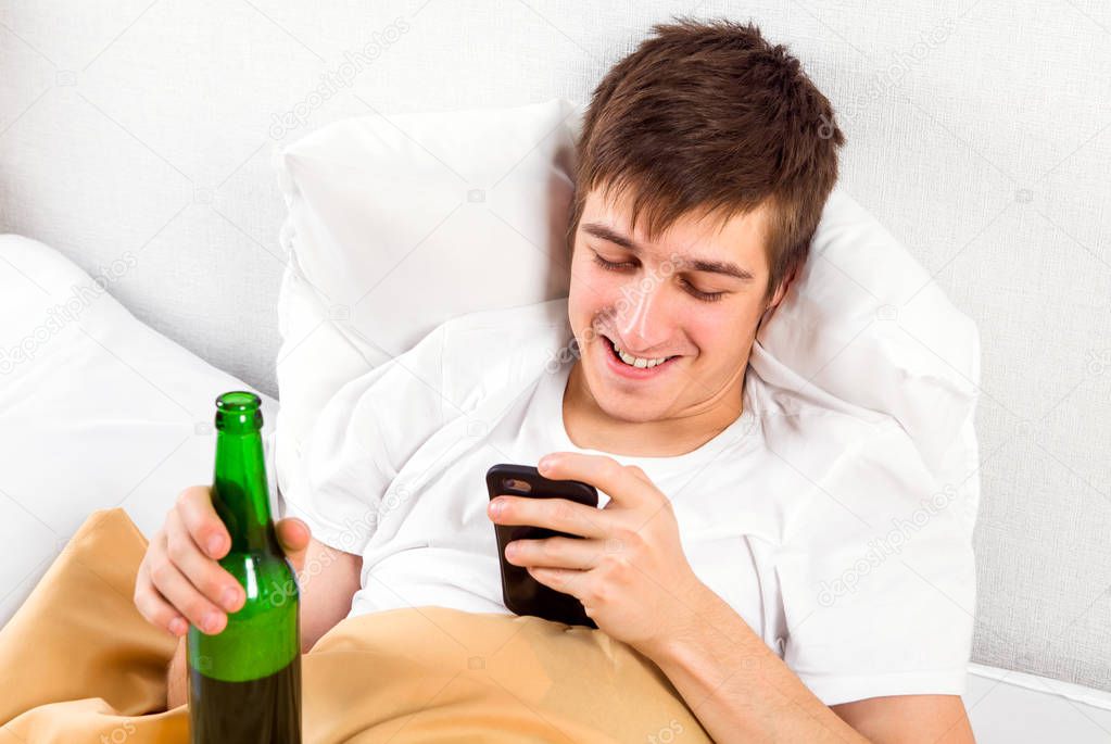 Cheerful Young Man with a Phone and a Beer Bottle in the Bed