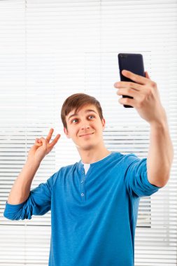 Mutlu Genç Adam, bir odanın arka planında cep telefonuyla selfie çekiyor.