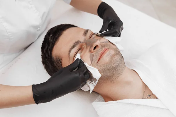 Kosmetikerin trägt im Wellness-Salon Kosmetikmaske auf das Gesicht des Mannes auf. — Stockfoto