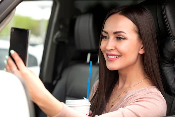 Retrato de motorista menina amigável muito sorridente fazendo uma selfie em um carro com um copo de papel — Fotografia de Stock