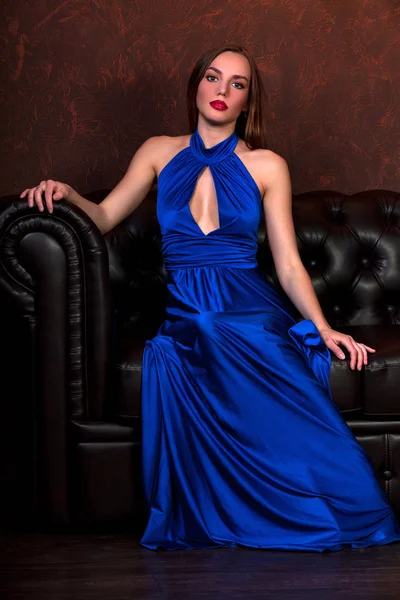 Uzun mavi elbisesi bir deri kanepede oturan güzel kadın — Stok fotoğraf