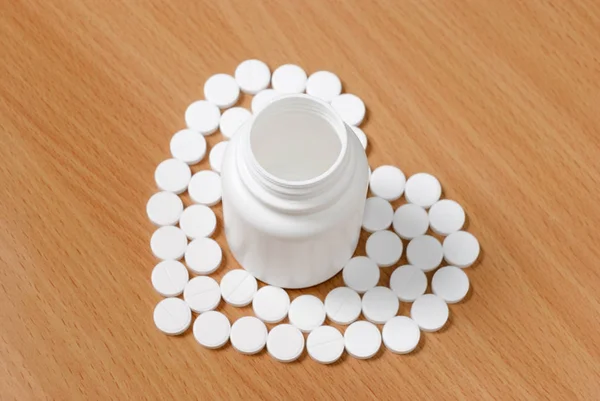 White pills and bottle on flat polished wooden surface. Pills arranged in heart around the bottle. Pills and bottles are white. Bottle is opened.