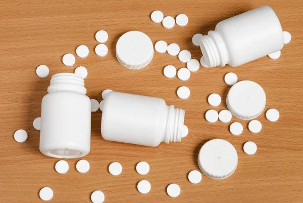 White pills and bottles scattered on flat polished wooden surface. Pills and bottles are white. Bottles are opened. Covers lay nearby.