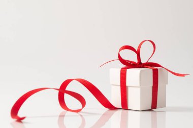 Üzerinde kırmızı saten bir kurdele bulunan küçük bir kare beyaz hediye kutusu Noel, yeni yıl ya da doğum günü teması için mükemmel bir fiyonk oluşturuyor. Kurdele, kutunun etrafında yansıtıcı beyaz bir yüzey etrafında dönüyor.