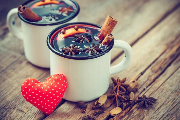 Mulled wine in mugs, red heart and spice on old wooden table.