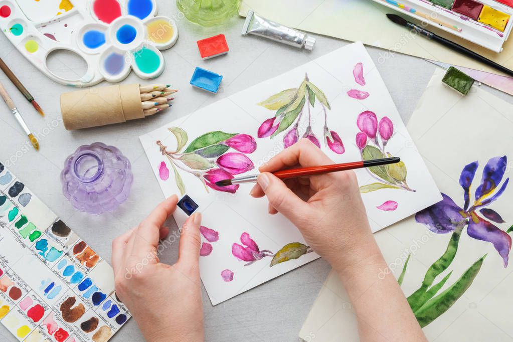 Watercolor drawing - pink cherry flowers - and artistic equipment on desk. Top view. Painter drawing at working place.