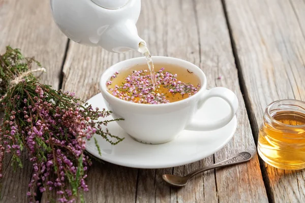 Té de brezo saludable vertido en taza blanca. Tetera, tarro de miel pequeña y racimo de brezo . —  Fotos de Stock