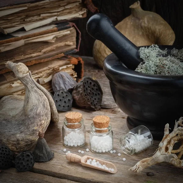 Flessen van homeopathische bolletjes, mortel, gedroogd mos, oude boeken, droge wortels, noten en planten op tafel. Homeopathie geneeskunde. — Stockfoto