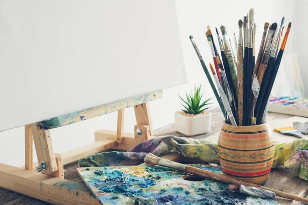 Artistic equipment in studio: canvas on wooden easel, paint brushes, paints and used palette. — Stock Photo, Image