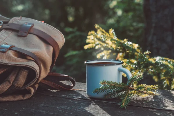 Enameled cup of coffee or tea, backpack of traveller on wooden board in summer forest outdoors. — Stock Photo, Image