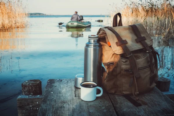 Emaljerad mugg med kaffe eller te, ryggsäck med resenär och termos på träbrygga på lugn sjö. En fiskare på gummibåt i bakgrunden. — Stockfoto