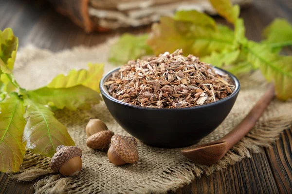 Healthy oak bark in black ceramic bowl, accorns, green oak leaves and old book on background. Alternative medicine.