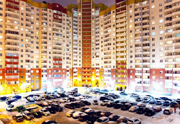 Multi-family house at night with many light windows and yard with parking with cars