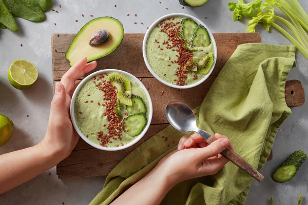 Breakfast detox green smoothie bowl with fresh kiwi, flaxseeds, cucumber and avocado in the female hands on a wooden board with a green napkin and celery sprigs, top view. Super Food