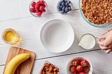 Kahvaltı hazırlarken, bir bardak süt ile beyaz ahşap bir masa üzerinde tutan bir kadın eli boş plaka, sulu çilek, bal, muz, granola ve fındık. Sağlıklı gıda. Adım adım tarif fotoğraf. Üstten görünüm