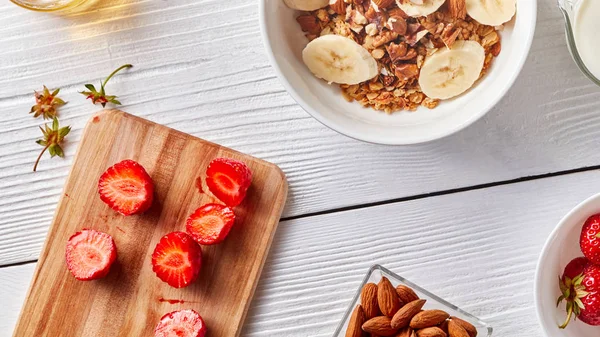 Beyaz ahşap bir masa kopya alanı, bir ahşap tahta ve badem bir plaka üzerinde çilek parçaları ile organik granola ve muz dilimleri ile plaka. Sağlıklı bir kahvaltı için organik malzemeler. Adım adım pişirme