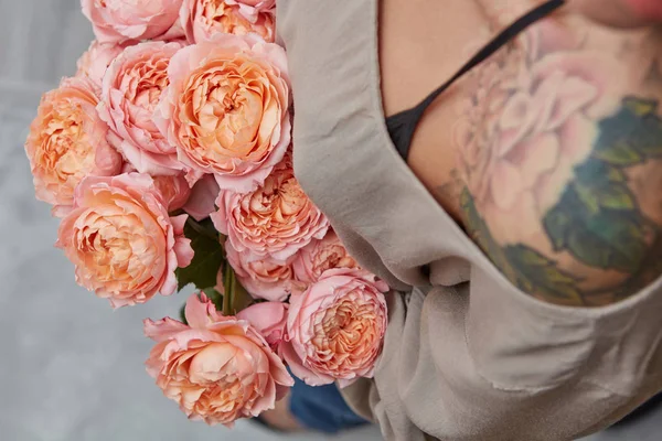 一个女人在她的肩膀上有一个纹身 是在灰色的背景上持有微妙的明亮的粉红色玫瑰 情人节礼物的德托普观 — 图库照片
