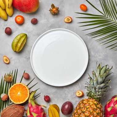 Boş bir beyaz plaka bileşimi yeşil palmiye yaprakları, ananas, mango, carambola, lychee, mandarin ve tutku meyve gri mermer tablo üzerinde bir kopya alanı ile. Düz yatıyordu
