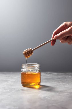 Kadın elini tut çiçek bal damlayan bir cam kavanoz üzerinde gri bir arka plan, metin için yer ile tahta bir sopa. Saf organik tatlı iyilik.