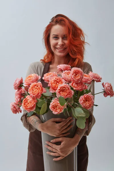 一个面带微笑的红头发的女人在棕色围裙举行了一个花瓶与玫瑰在一个年的颜色2019年生活珊瑚潘通在灰色的背景与文本的空间 花店的概念 — 图库照片