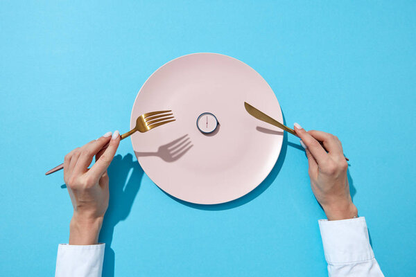 Round watch of six o'clock and woman's hand with fork and knife in girl's hands on a blue background with shadows. Time to lose weight, eating control or diet concept. Place for text.