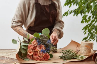 Kahverengi apron içinde çiçekçi kız hafif bir arka planda taze gül mercan buket yaratıyor. Adımda adım işlem. Çiçek teslimatı ile küçük iş. Anneler günü.