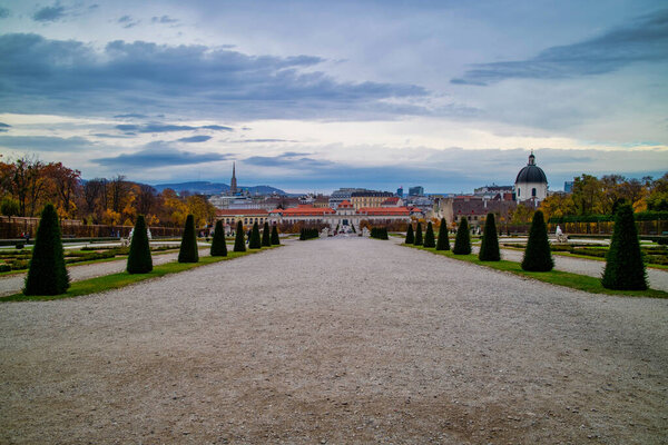 Прекрасный пейзаж с широкой главной пешеходной аллеей перед Unteres Belvedere с регулярной посадкой деревьев и растений в Вене, Австрия на фоне облачного неба.