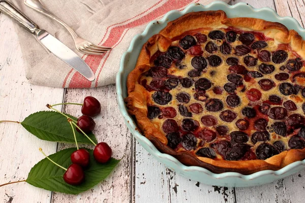 Homemade cherry tart in blue baking pan with raw red cherries, linen kitchen towel, fork and knife on rustic table