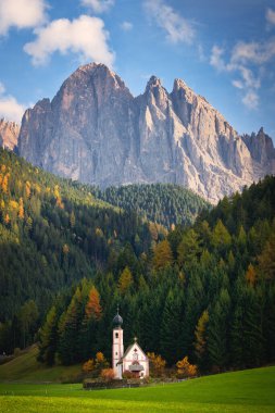 The small church of San Giovanni in Ranui or Sankt Johann in Ranui in the grass with behind it a forest with larch trees in autumn colors and further behind the mighty Geisler Dolomites mountain peak in a landscape in Villnoess in South Tyrol, Italy. clipart