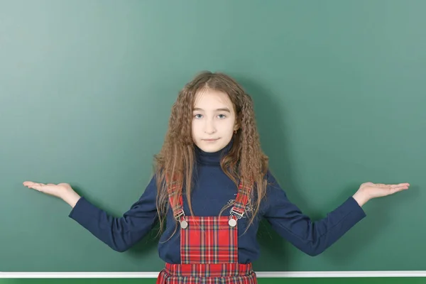 School meisje in de buurt van groene schoolboard. Glimlachend klein school meisje fokken armen aan de zijkanten op groene schoolboard. — Stockfoto