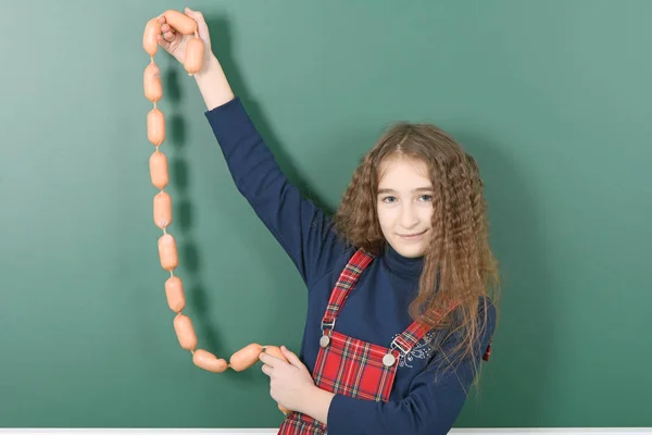 Colegiala cerca de la junta escolar verde. Joven chica juguetona sostiene la cadena de salchichas en el tablero de la escuela verde . —  Fotos de Stock