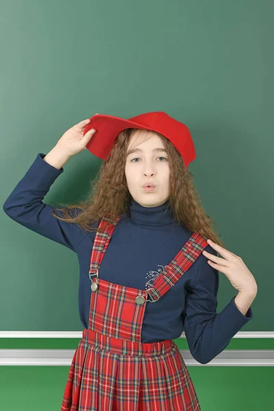 Colegiala cerca de la junta escolar verde. Joven juguetona chica sostiene una gorra roja . — Foto de Stock