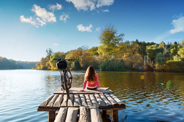 Ragazza siede sulla riva del fiume. bici sul fiume con una borsa — Foto Stock