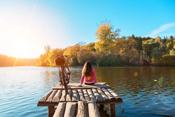 Chica se sienta en la orilla del río. bicicleta en el río con una bolsa —  Fotos de Stock