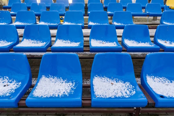 Stadium seats in the snow in winter. Snow-covered blue plastic seats at the small stadium in winter. Snowfall interferes with sports. Uncleaned stadium.