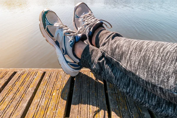 Man with crossed legs relaxing on the wooden pier. Young guy wearing sneakers sitting by the lake in summer afternoon.