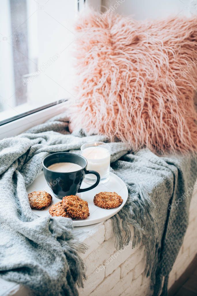 Scandinavian style breakfast, cup of coffee and cookies on cozy windowsill with warm blanket and pillow