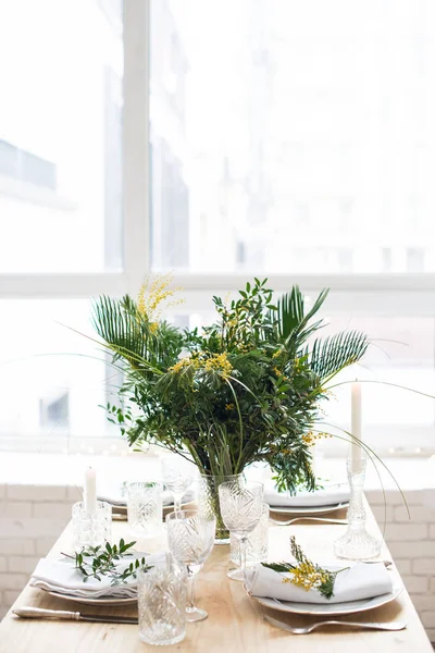 Beautiful springtime table setting with green leaves and mimosa branches, bright white table dinner decoration