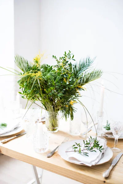 Beautiful springtime table setting with green leaves and mimosa branches, bright white table dinner decoration