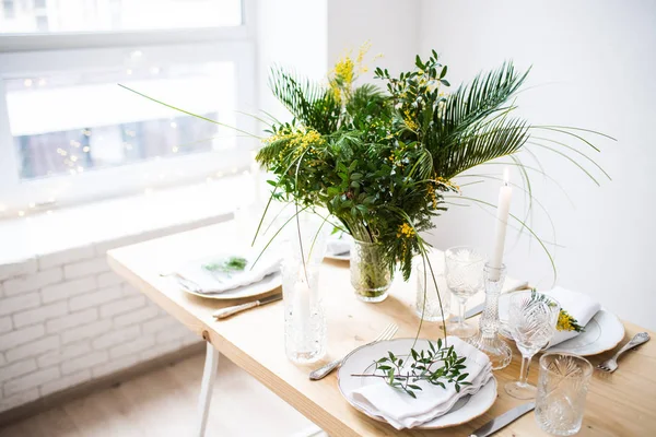 Beautiful springtime table setting with green leaves and mimosa branches, bright white table dinner decoration